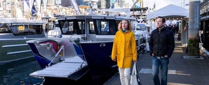 Two people walk along a marina and look at docked boats.