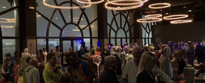 A lively costume party scene with colorful outfits, chandeliers, and guests mingling in a spacious venue, illuminated by soft lighting.