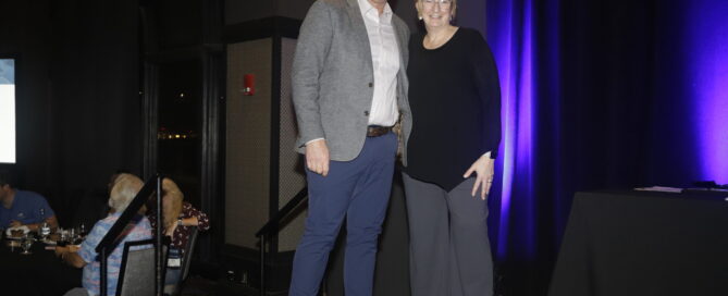 An award winner and a presenter stand on stage, smiling together. The backdrop features purple lighting, with tables set for an event visible behind them.