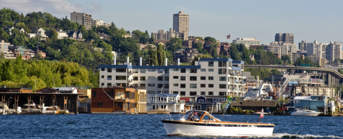 Wood motorboat navigating a harbor, dotted with houseboats and backed by hills and city buildings.