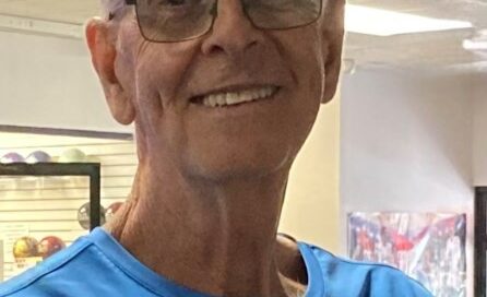 Tom Hunter, smiling and wearing glasses and a blue shirt, standing indoors with colorful bowling equipment visible in the background.