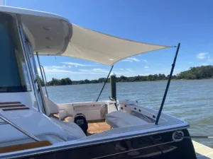 View from a boat featuring a large white canopy, wooden seating area, and calm water with blue sky and greenery in the background.