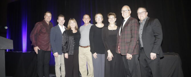Group of eight members of the MFA advisory board posing together on stage, dressed in formal attire against a dark backdrop with purple lighting.