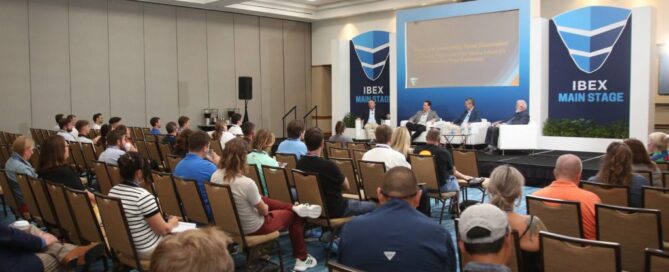 A conference panel discussion in a spacious venue, featuring speakers on stage with an audience seated in rows, and blue decorative elements.