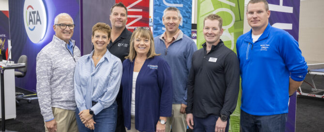 A_group of seven smiling professionals stands in front of vibrant banners, showcasing a mix of blue, green, and red colors at an event.